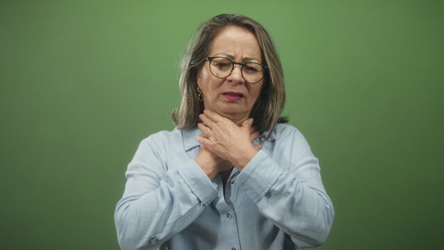 Hispanic woman with grey hair clutching her throat with both hands in a green studio setting, eyes closed and mouth open showing choking distress; panic medical concern.