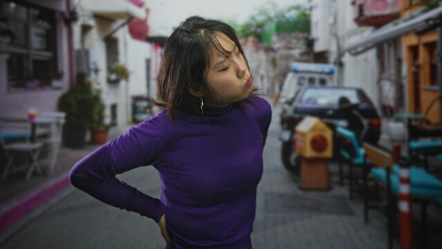 Woman holds lower back with hand, head tilted upward and eyes closed on a narrow street near cafe tables and parked scooter, purple sweater visible, backache; discomfort relief.