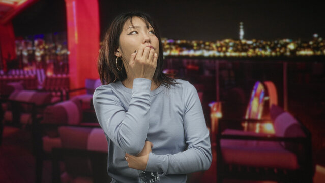 Woman young beautiful holding a water bottle, hand to mouth gesture with elbow tucked across torso in rooftop building lounge overlooking city lights at night; pensive solitude.