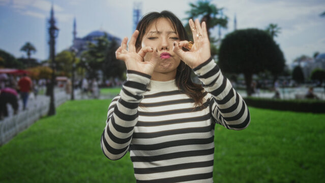 Young woman in striped sweater makes ok gesture with both hands and puckered lips while standing on green lawn in front of historic building; playful approval.