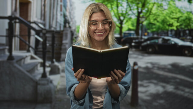 Woman holding open book with both hands, smiling and reading with expressive gestures on a street by building steps and parked cars, wearing glasses and denim shirt; joyful.