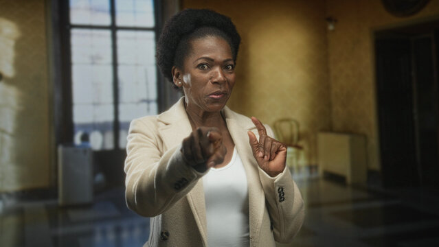 Woman points finger, hands visible, wearing beige coat and white top in a building; confidence encouragement warmth.