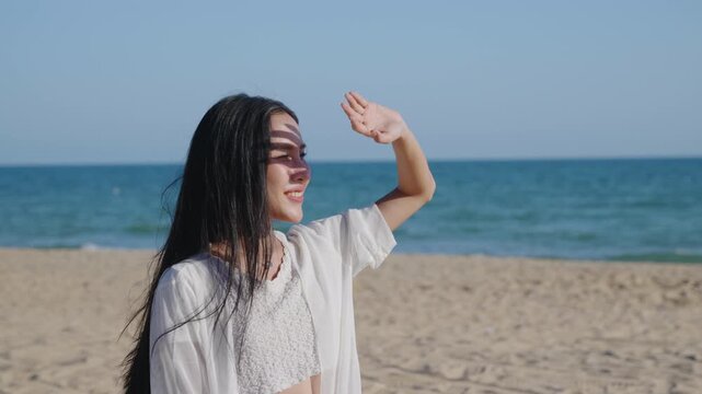During summer vacation on beach, smiling woman shielding eyes from sun, looking away while enjoying beautiful tropical sea landscape, feeling happy and relaxed on warm sunny day.