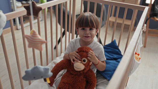 Boy in crib holds a brown stuffed monkey and reaches hand toward a hanging star mobile inside a building nursery, seated on white mattress with blue pillow; playful curiosity.
