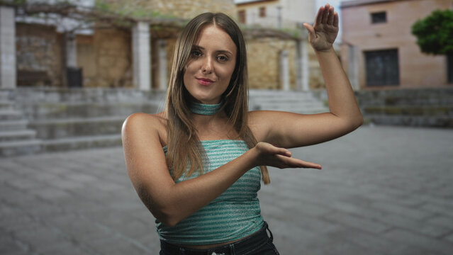 Young brunette woman with bare arms presenting palms as if holding an invisible object, posing in a stone plaza in front of a building; curiosity.