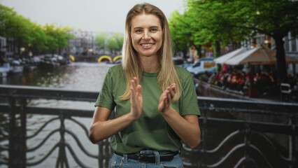 Fototapeta premium Woman clapping hands on a canal bridge by a street cafe with trees and water in view; celebration joy.