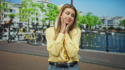 Fototapeta premium Blonde woman wearing visitor card holds hands to cheeks and gestures on a street by a canal; thoughtful.