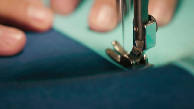 Close-up footage of a sewing machine needle and presser foot actively stitching dark blue and teal fabric, with fingers carefully guiding the material along the seam.