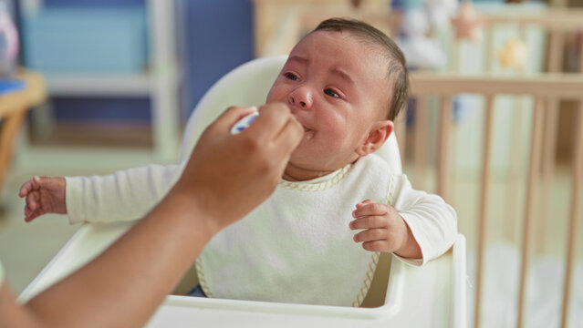 Baby boy crying and spitting spoonful of puree into mouth with messy bib while seated in highchair in building; feeding frustration.