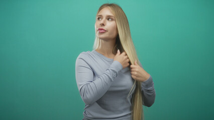 Fototapeta premium Woman touching long blonde hair and shoulder while smoothing sleeve in a studio with teal backdrop and soft gaze; contemplation.