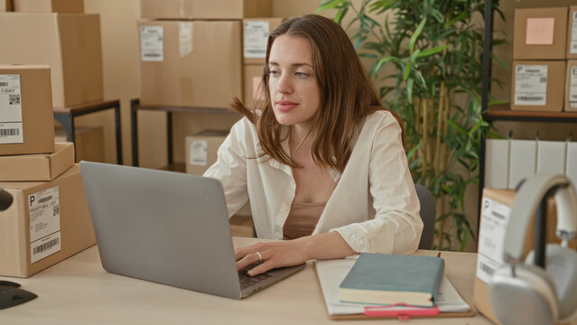 Woman typing on laptop with hands on keyboard amid stacked parcels and shipping labels at a packing table in a building; small business optimism.