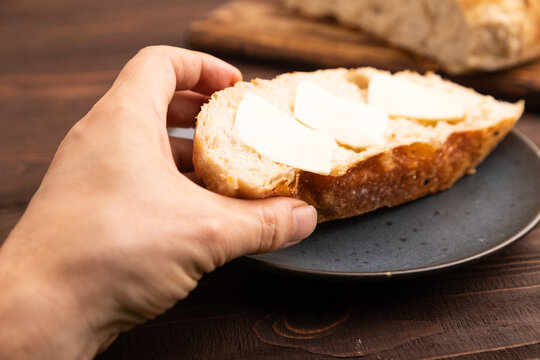 Slice of homemade bread with butter on plate with hand on brown wooden, close up, side view, selective focus.