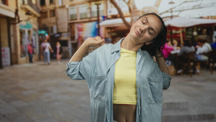 Fototapeta premium Woman pulls denim shirt collar and stretches shoulders with eyes closed in a busy street cafe; relaxed vacation.