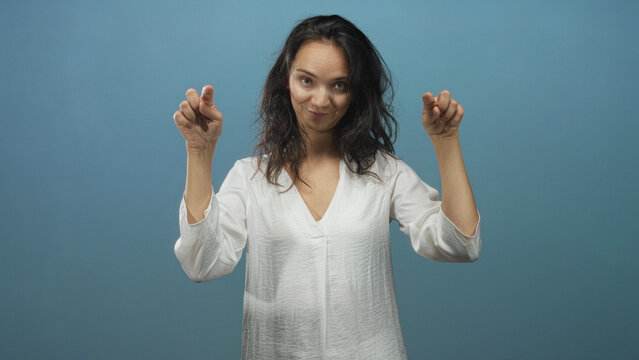 Woman in white blouse pointing fingers down with puckered lips and closed eyes against blue studio backdrop; playfulness.