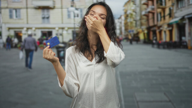 Woman holding blue creditcard, hand covering mouth with closed eyes on a busy street lined with cafes; financial worry surprise.