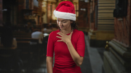 Fototapeta premium Woman with hand on chest touching heart, wearing santa hat and red top in a church building, looking down; quiet reflection.