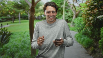 Fototapeta premium Man in gray sweater taps smartphone with right index finger while smiling on a tree lined path in forest; relaxed connection.