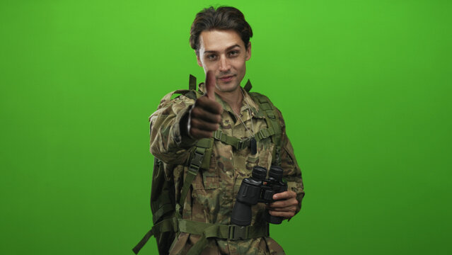 Soldier in camouflage uniform wearing backpack and dog tags, holding binoculars and giving thumbs up to camera in studio; approval duty morale.