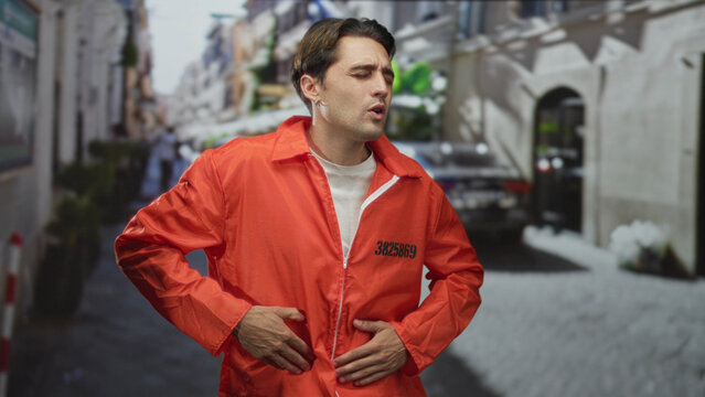 Man in orange jumpsuit clutching his stomach with hands over zipper and inmate number visible on a street lined with bollards and plants; suffering pain.