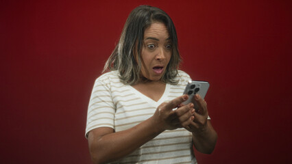 Woman middle aged holding smartphone with both hands, eyes wide and hand covering mouth while...