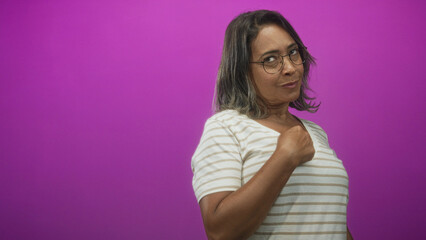 Obraz premium Hispanic middle age woman points finger to chest, wearing striped shirt and glasses against a magenta studio backdrop and soft lighting; confidence affirmation.