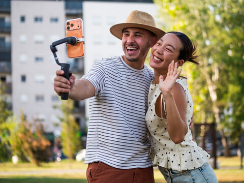 Cheerful couple embracing and creating joyful social media content with a smartphone stabilizer in a sunny park.