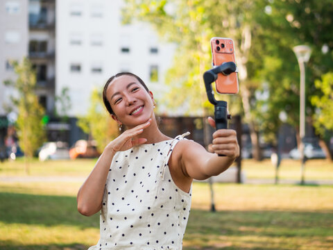 Smiling content creator filming herself with a smartphone stabilizer during a sunny day in the park.