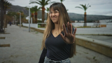 Fototapeta premium Young woman wearing glasses with outstretched hand making a stop gesture toward camera on a street promenade lined with palm trees and benches; playful refusal.