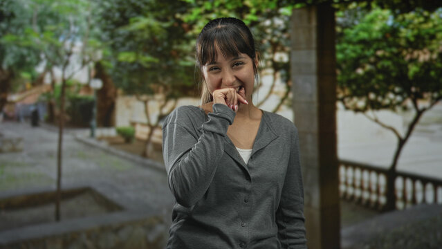 Young woman biting her hand and smiling, fingernails visible, wearing gray cardigan, standing on a street with trees and stone balustrade; playful shyness.