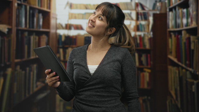 Young hispanic woman holding tablet and tapping screen with index finger while smiling and looking at the display in library; curiosity learning.