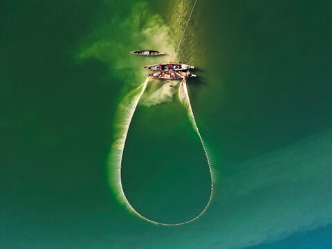 Rangamati, Chittagong Division, Bangladesh - 14 December 2024: Aerial view of traditional wooden fishing boats casting a large circular net into the clear green waters of Kaptai Lake.