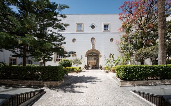 The lush inner garden courtyard and white facade with architectural windows at the Royal Palace of Palermo in Sicily.