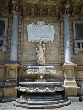 Vertical image of the 17th-century stone facade at Piazza Vigliena in Palermo, featuring a Latin dedication plaque above an ornate marble fountain and statue.