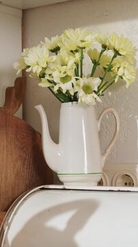 Close up of white ceramic pitcher filled with yellow daisies on shelf. Wooden cutting board and old enamelware object are. Electrical outlet is partially seen below scene