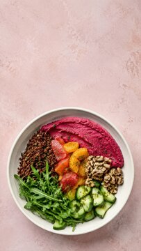 Vegan bowl featuring quinoa, colorful vegetables, beet hummus, and arugula arranged in a circular white dish on a textured pink background