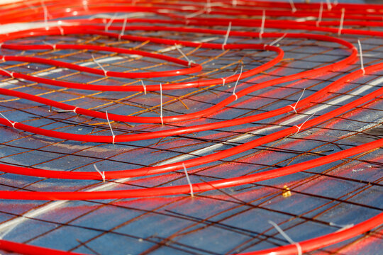 Red tubing lies coiled on a metal grid. Metal grid provides structural support beneath the tubes. Tubes secured with small white fasteners. Setup ready for heating system installation.