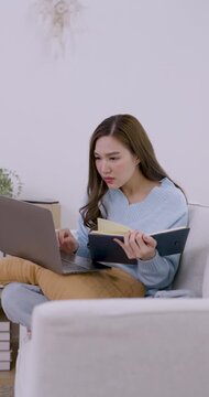 Young Asian woman sits on sofa with laptop and smartphone, checks screen, switches mobile device while doing remote work inside cozy minimal apartment vertical video today indoors now relaxed