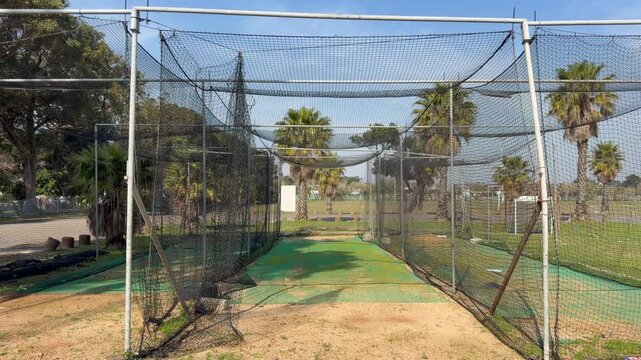 : A tunnel-like cricket practice net stretches toward an open sports field, framed by black netting and white metal posts. Green synthetic turf runs down the center of the sandy enclosure.
