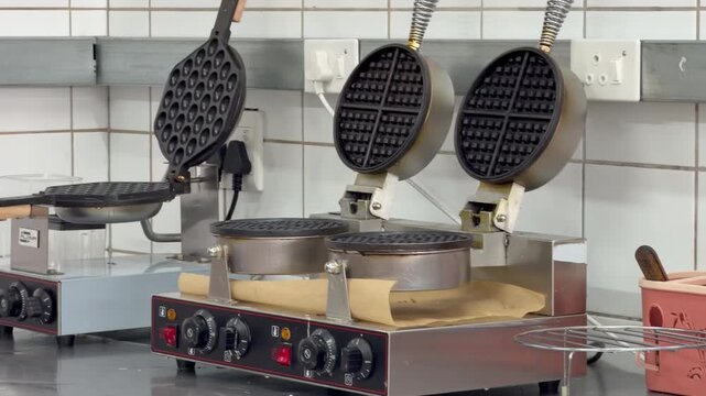 Close-up view of commercial waffle-making equipment in a professional kitchen. Multiple electric waffle irons and cooking appliances are arranged on a stainless-steel countertop.
