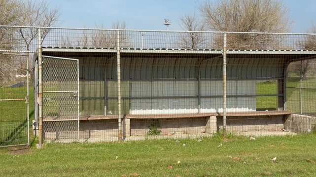 A long, covered dugout with a curved metal roof sits behind protective chain-link fencing on a quiet baseball field.