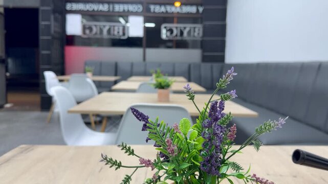 Cinematic interior shot of a modern contemporary restaurant or caf&eacute; with neatly arranged empty dining tables, white chairs, grey upholstered seating, and decorative purple and green flowers.