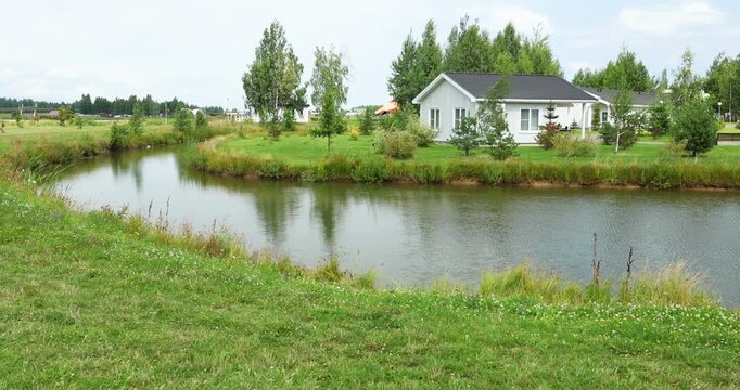 Beautiful landscape design featuring winding river bend next to cozy cottage. Scenic waterfront property surrounded by manicured green lawn and young trees. Peaceful countryside location.