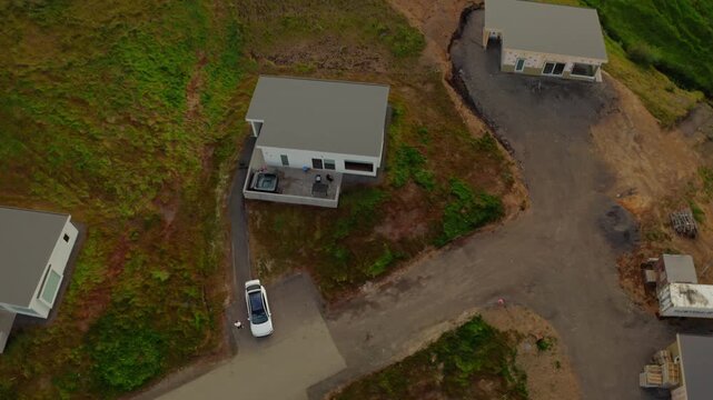 An aerial drone shot flies over a modern white house with a flat roof and a hot tub on a hillside in Akureyri, Iceland. Another house is under construction nearby.