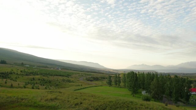 An aerial shot flies over the green, rolling hills and countryside near Akureyri, Iceland. A fjord and hazy mountains are visible in the distance under a cloudy sky.
