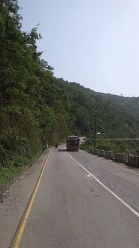 Vertical footage of a scenic paved highway winding through lush green mountain terrain in Nepal. The road passes between densely vegetated hills with utility lines and roadside safety barriers