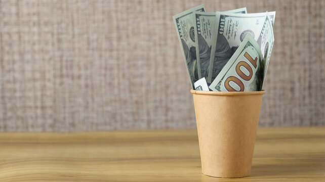 Money bills placed in a brown paper cup on a wooden table against a neutral background