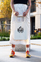 A young woman stands outdoors holding a tote bag with a leaf design. She wears a white skirt and colorful sneakers. The background features greenery and modern architecture. © Nadezhda Zaitceva