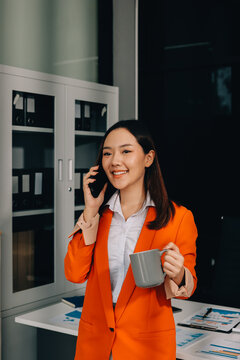 Smiling asian businesswoman talking on phone and using laptop while working at office desk