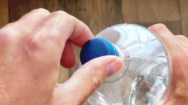 Top view close up shot of hands opening a clear plastic water bottle with a blue screw cap against a wooden surface background. Hydration concept.