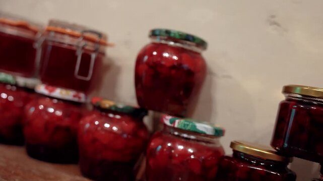 Rows of glass jars filled with red homemade preserves and fruit compote arranged on a rustic wooden shelf, with shallow depth of field and warm indoor lighting